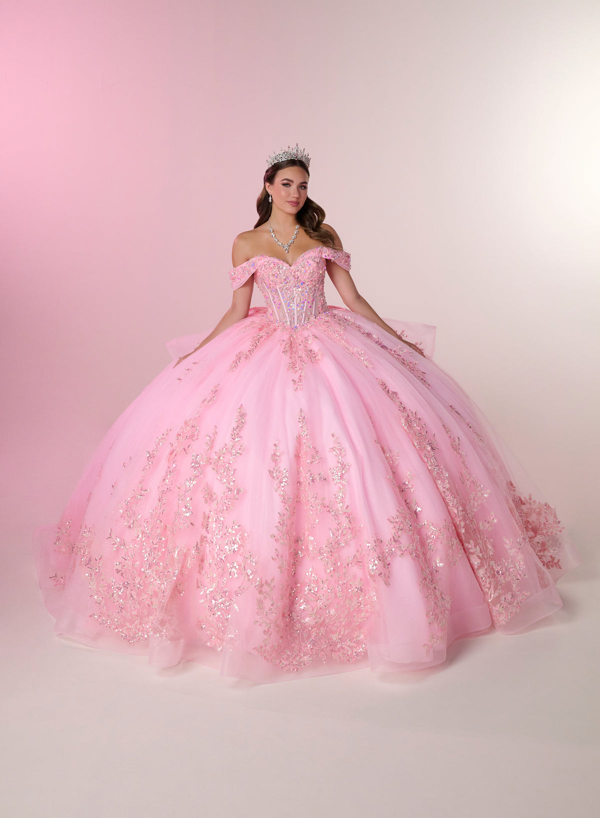 A young woman wearing a voluminous light pink ball gown with off-the-shoulder straps, a fitted embellished bodice, and a full tulle skirt decorated with floral lace and sequins, posed against a soft pink studio backdrop. / Stunning off-shoulder ball gown with shimmering embellished appliqués, a structured corset bodice, a full tulle skirt, and a dramatic oversized bow with a sweeping train.
