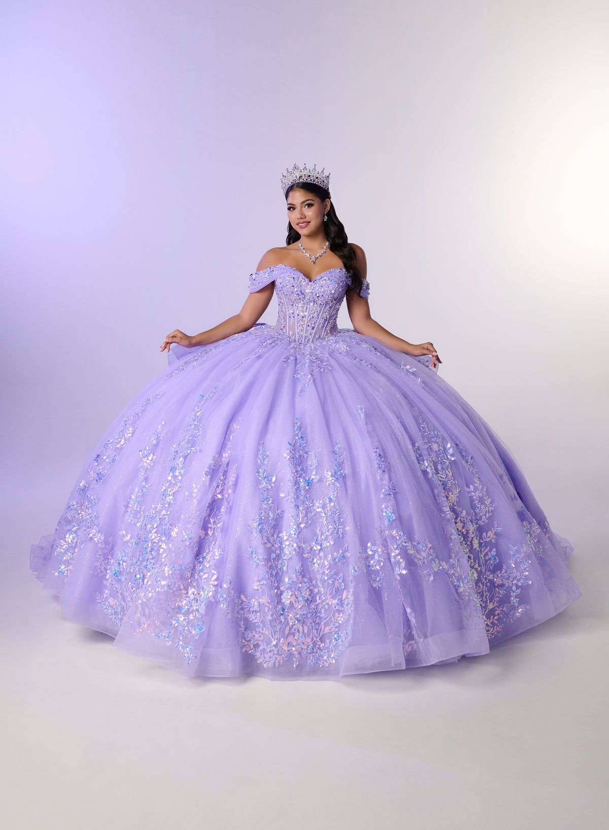 A woman wearing a voluminous lavender quinceañera ball gown with off-the-shoulder straps, a sweetheart neckline, and iridescent floral embroidery, posed against a light lavender studio backdrop with a tiara. / Stunning off-shoulder ball gown with shimmering embellished appliqués, a structured corset bodice, a full tulle skirt, and a dramatic oversized bow with a sweeping train.