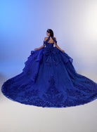 A woman wearing a voluminous royal blue ball gown with intricate lace and bead embellishments, photographed from behind against a blue-to-white studio backdrop. / Elegant ball gown featuring a beaded off-shoulder bodice, cascading ruffle details, intricate embellishments, and a dramatic oversized bow with a sweeping train.