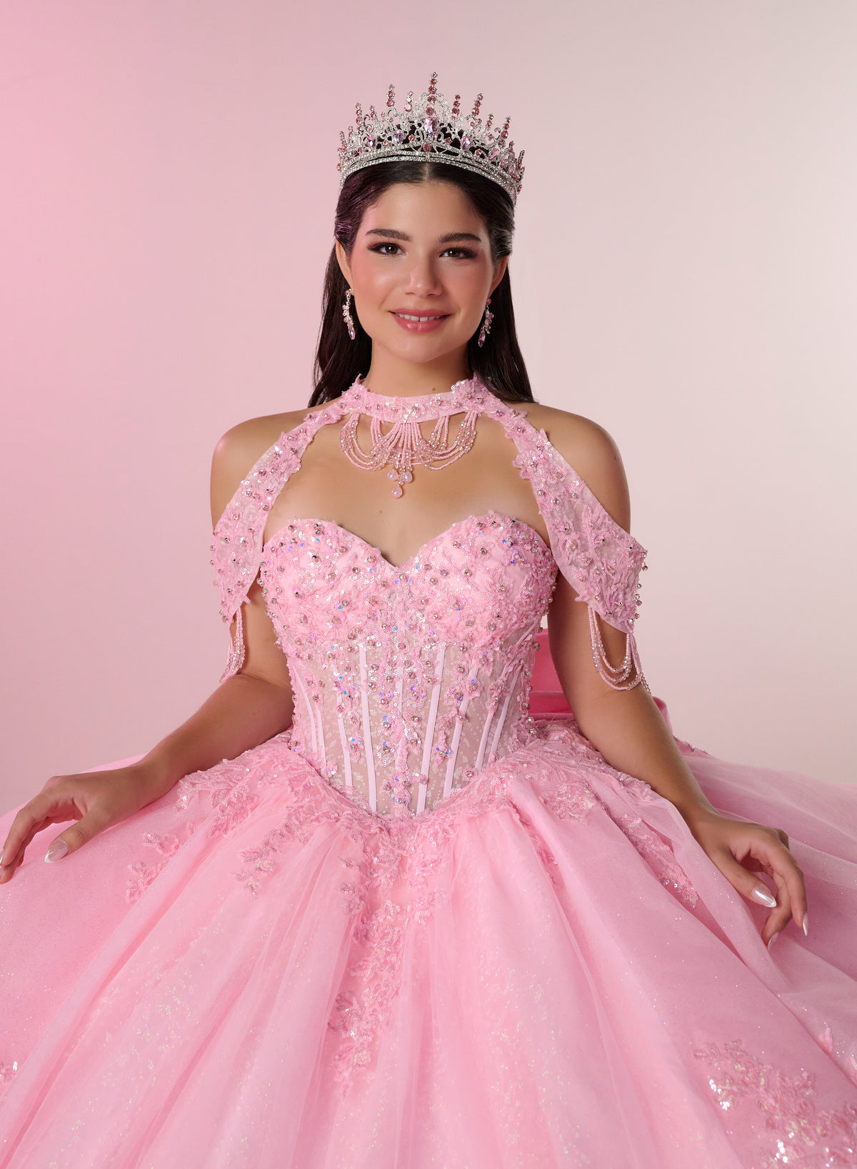 A woman wearing an ornate pink ball gown with a corset-style bodice, cold-shoulder lace sleeves, and a full tulle skirt, posed against a soft pink studio background. She is also wearing a jeweled tiara and dangling earrings. / Elegant ball gown featuring a beaded off-shoulder bodice, cascading ruffle details, intricate embellishments, and a dramatic oversized bow with a sweeping train.