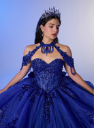 A woman wearing an elaborate royal blue quinceañera ball gown with off-the-shoulder straps, a beaded corset bodice, and a voluminous tulle skirt, styled with a matching jeweled tiara against a soft blue studio background. / Elegant ball gown featuring a beaded off-shoulder bodice, cascading ruffle details, intricate embellishments, and a dramatic oversized bow with a sweeping train.