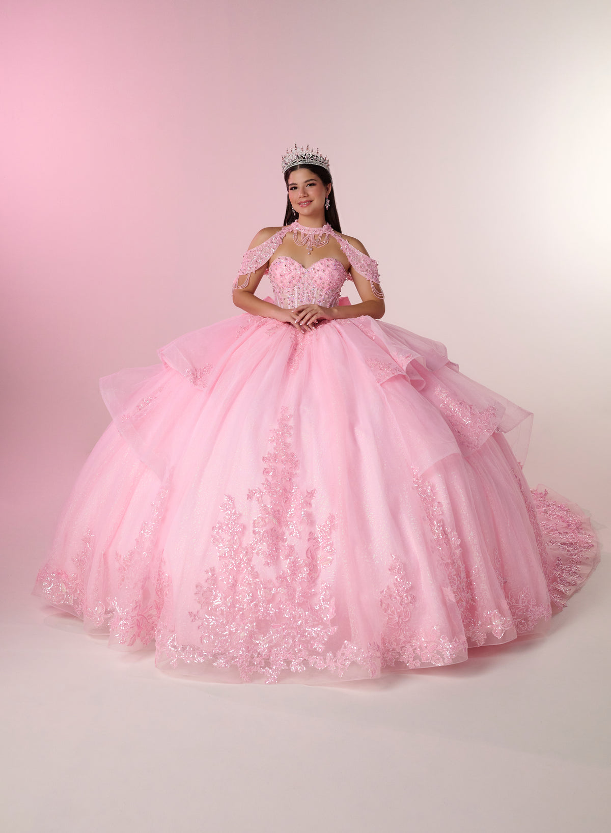 A young woman wearing a voluminous light pink ball gown with lace applique and a tiara, standing against a soft pink studio backdrop. / Elegant ball gown featuring a beaded off-shoulder bodice, cascading ruffle details, intricate embellishments, and a dramatic oversized bow with a sweeping train.