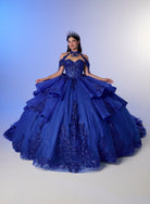 A woman wearing a voluminous royal blue ball gown with lace applique and a matching choker and tiara, posed against a soft blue-to-white studio backdrop. / Elegant ball gown featuring a beaded off-shoulder bodice, cascading ruffle details, intricate embellishments, and a dramatic oversized bow with a sweeping train.