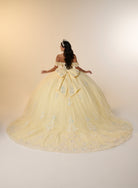 A woman stands with her back to the camera wearing an off-the-shoulder pale yellow ball gown with a large bow at the back and light blue floral embroidery, set against a plain beige studio background. / Romantic off-shoulder ball gown with delicate embellished appliqués, a fitted bodice, a voluminous tulle skirt, and a statement bow at the back.