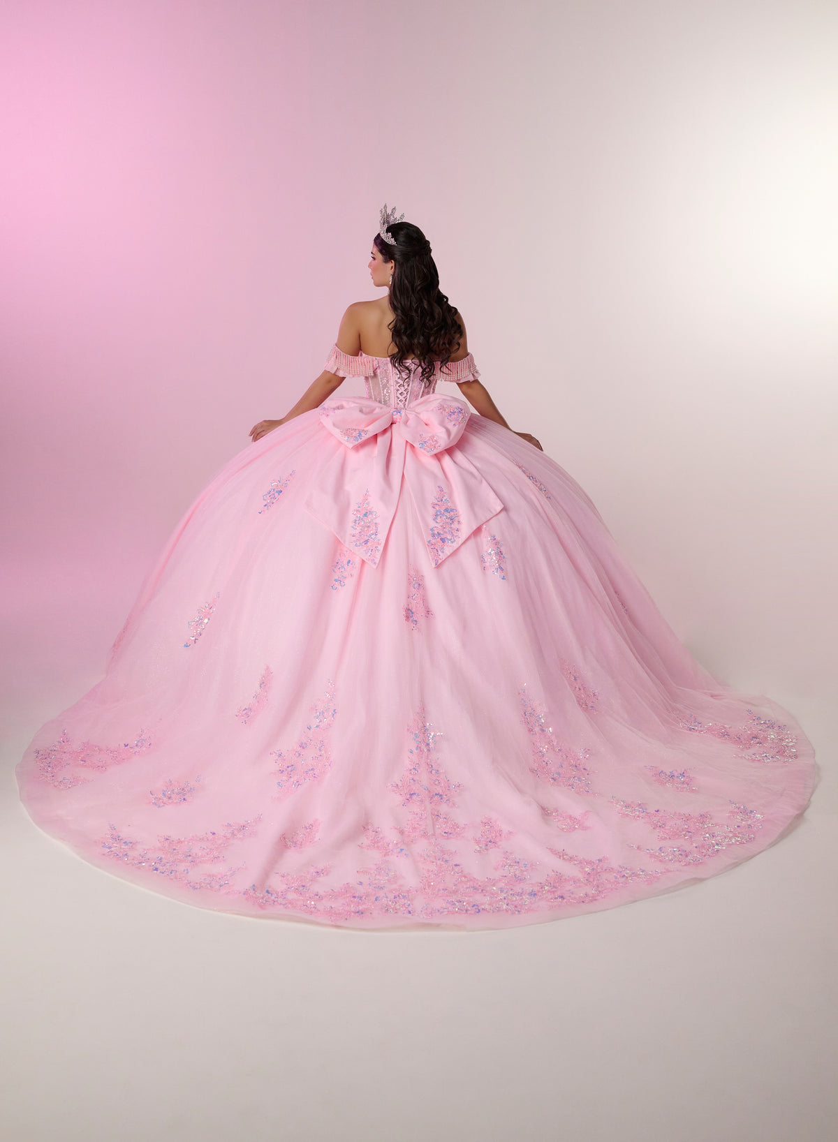 A woman wearing a voluminous light pink ball gown with floral embroidery and a large bow at the back, photographed from behind against a soft pink-to-white studio backdrop. / Romantic off-shoulder ball gown with delicate embellished appliqués, a fitted bodice, a voluminous tulle skirt, and a statement bow at the back.