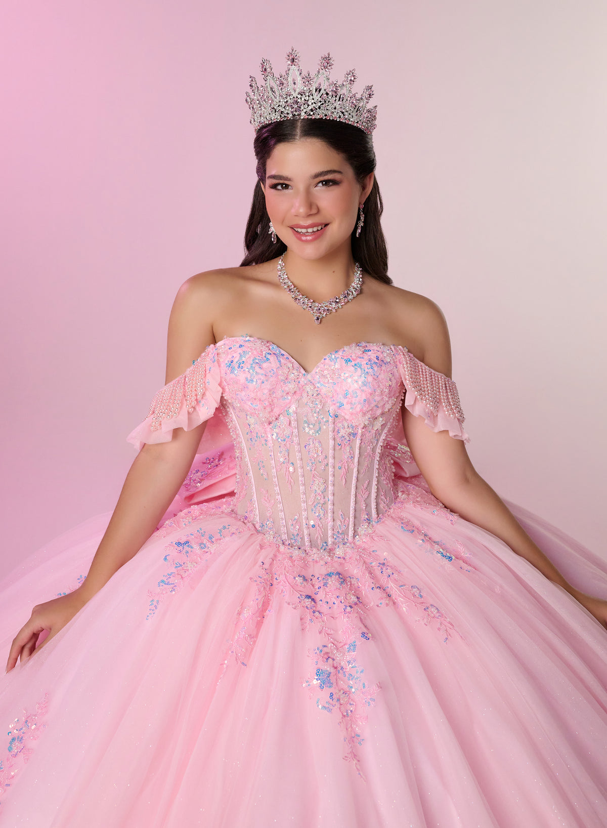A young woman wearing a sparkling off-the-shoulder pink quinceañera ball gown with blue floral embroidery, a silver tiara, and a matching silver necklace, posed against a soft pink studio backdrop. / Romantic off-shoulder ball gown with delicate embellished appliqués, a fitted bodice, a voluminous tulle skirt, and a statement bow at the back.