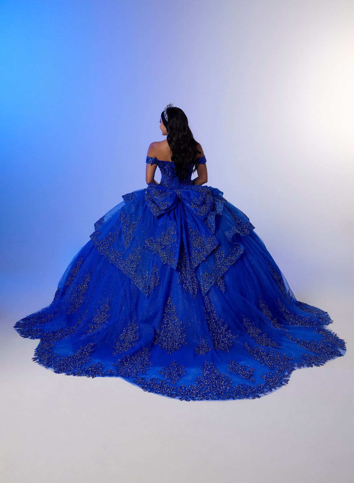 A woman viewed from behind wearing an elaborate royal blue ball gown with intricate lace and beadwork, set against a blue-to-white studio gradient background. / Luxurious off-shoulder ball gown featuring glittering embellishments, layered draped skirt details, and a dramatic cathedral-length train for a breathtaking princess-inspired look.