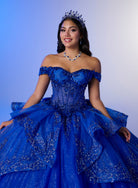 A woman wearing an elaborate royal blue off-the-shoulder ball gown with sparkling embellishments, a matching tiara, and a blue beaded necklace, posed against a blue-to-white gradient studio background. / Luxurious off-shoulder ball gown featuring glittering embellishments, layered draped skirt details, and a dramatic cathedral-length train for a breathtaking princess-inspired look.