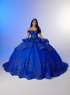 A woman wearing a voluminous royal blue off-the-shoulder ball gown with intricate silver floral embroidery, posed against a blue-to-white studio gradient background. / Luxurious off-shoulder ball gown featuring glittering embellishments, layered draped skirt details, and a dramatic cathedral-length train for a breathtaking princess-inspired look.