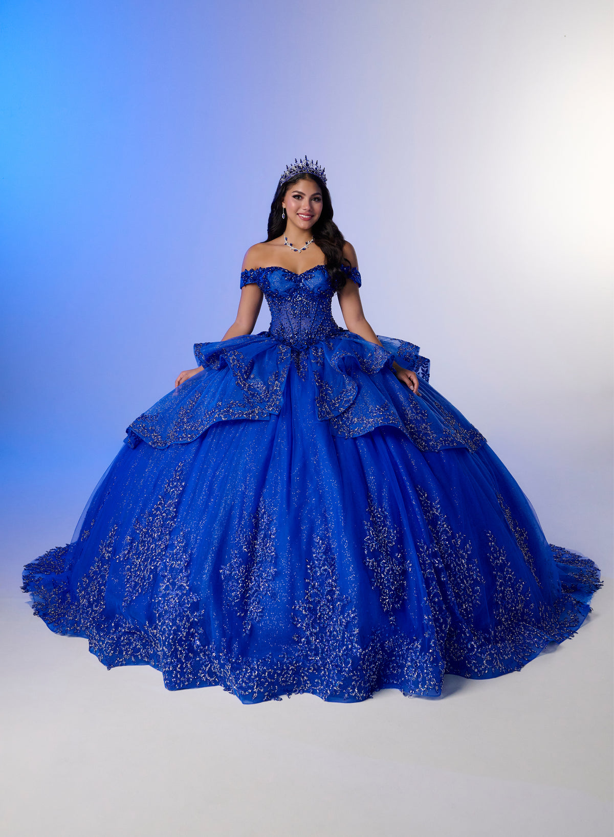 A woman wearing a voluminous royal blue off-the-shoulder ball gown with intricate silver floral embroidery, posed against a blue-to-white studio gradient background. / Luxurious off-shoulder ball gown featuring glittering embellishments, layered draped skirt details, and a dramatic cathedral-length train for a breathtaking princess-inspired look.