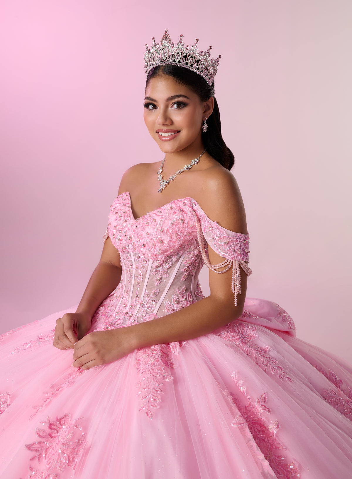 A young woman wearing a sparkling pink off-the-shoulder ball gown with lace applique, a silver tiara, and a matching silver necklace, posed against a soft pink studio background. / Glamorous off-shoulder ball gown with sparkling embellishments, a fitted corset bodice, a voluminous skirt, and a dramatic ruffled hem for a grand princess-inspired look.