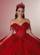 A smiling young woman wearing a voluminous red ball gown with off-the-shoulder straps, heavy beadwork, and a matching red tiara, photographed against a soft pink studio background. / Glamorous off-shoulder ball gown with sparkling embellishments, a fitted corset bodice, a voluminous skirt, and a dramatic ruffled hem for a grand princess-inspired look.