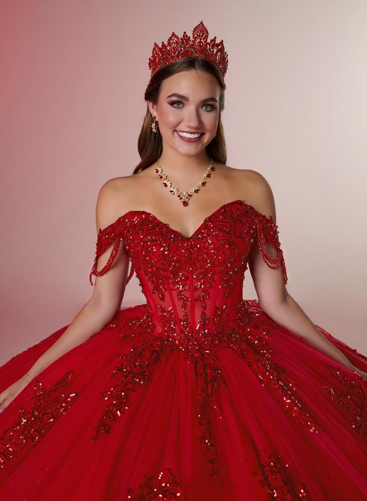 A smiling young woman wearing a voluminous red ball gown with off-the-shoulder straps, heavy beadwork, and a matching red tiara, photographed against a soft pink studio background. / Glamorous off-shoulder ball gown with sparkling embellishments, a fitted corset bodice, a voluminous skirt, and a dramatic ruffled hem for a grand princess-inspired look.