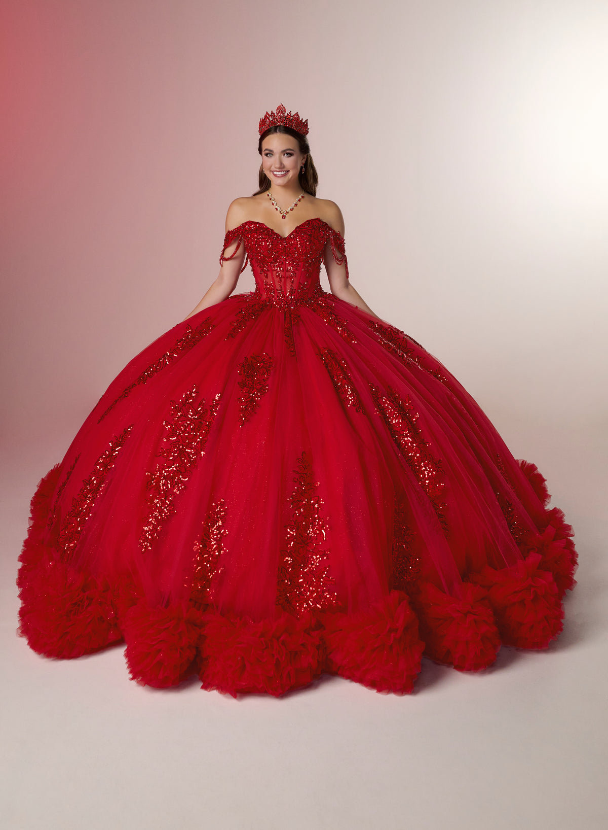 A woman wearing a voluminous red ball gown with gold sequin embellishments, a red tiara, and a red necklace, standing against a light beige studio background. / Glamorous off-shoulder ball gown with sparkling embellishments, a fitted corset bodice, a voluminous skirt, and a dramatic ruffled hem for a grand princess-inspired look.
