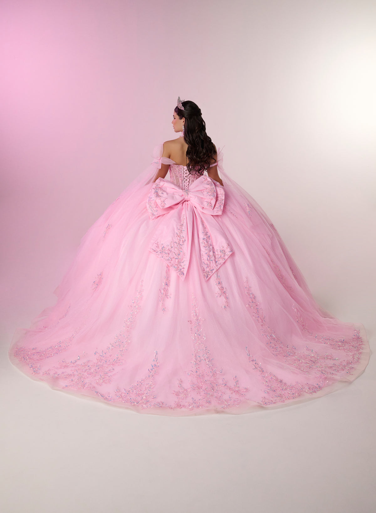 A woman viewed from behind wearing an elaborate off-the-shoulder pink ball gown with a very large bow at the back and embroidered tulle detailing, photographed against a soft pink-to-white studio background. / Dreamy off-shoulder ball gown featuring delicate sparkling appliqués, a structured corset bodice, a voluminous tulle skirt, and an oversized statement bow at the back.