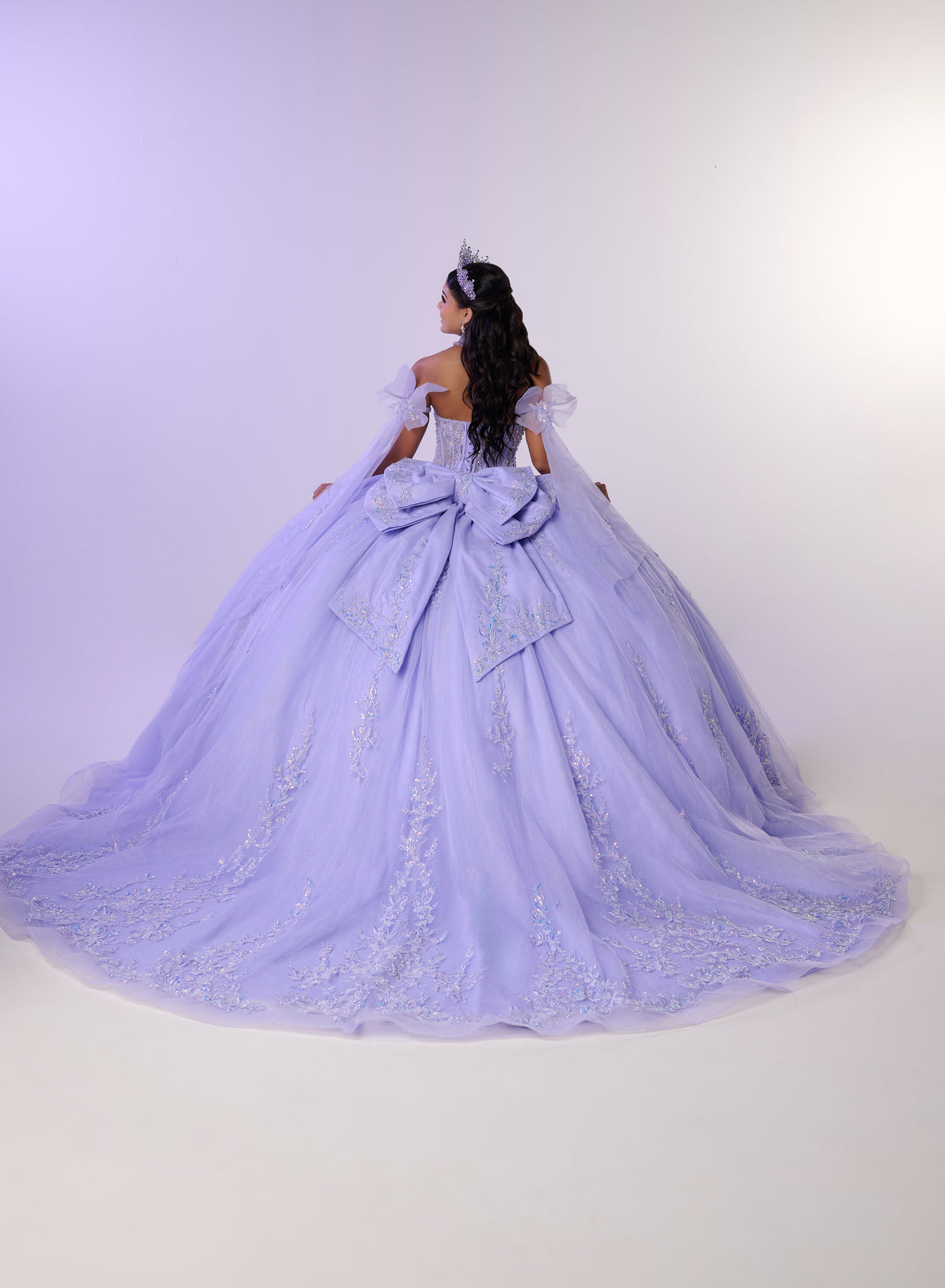 A woman photographed from behind wearing an elaborate lavender ball gown with off-the-shoulder straps, a large bow at the back, and a voluminous tulle skirt with embroidered detailing, against a plain light studio background. / Dreamy off-shoulder ball gown featuring delicate sparkling appliqués, a structured corset bodice, a voluminous tulle skirt, and an oversized statement bow at the back.
