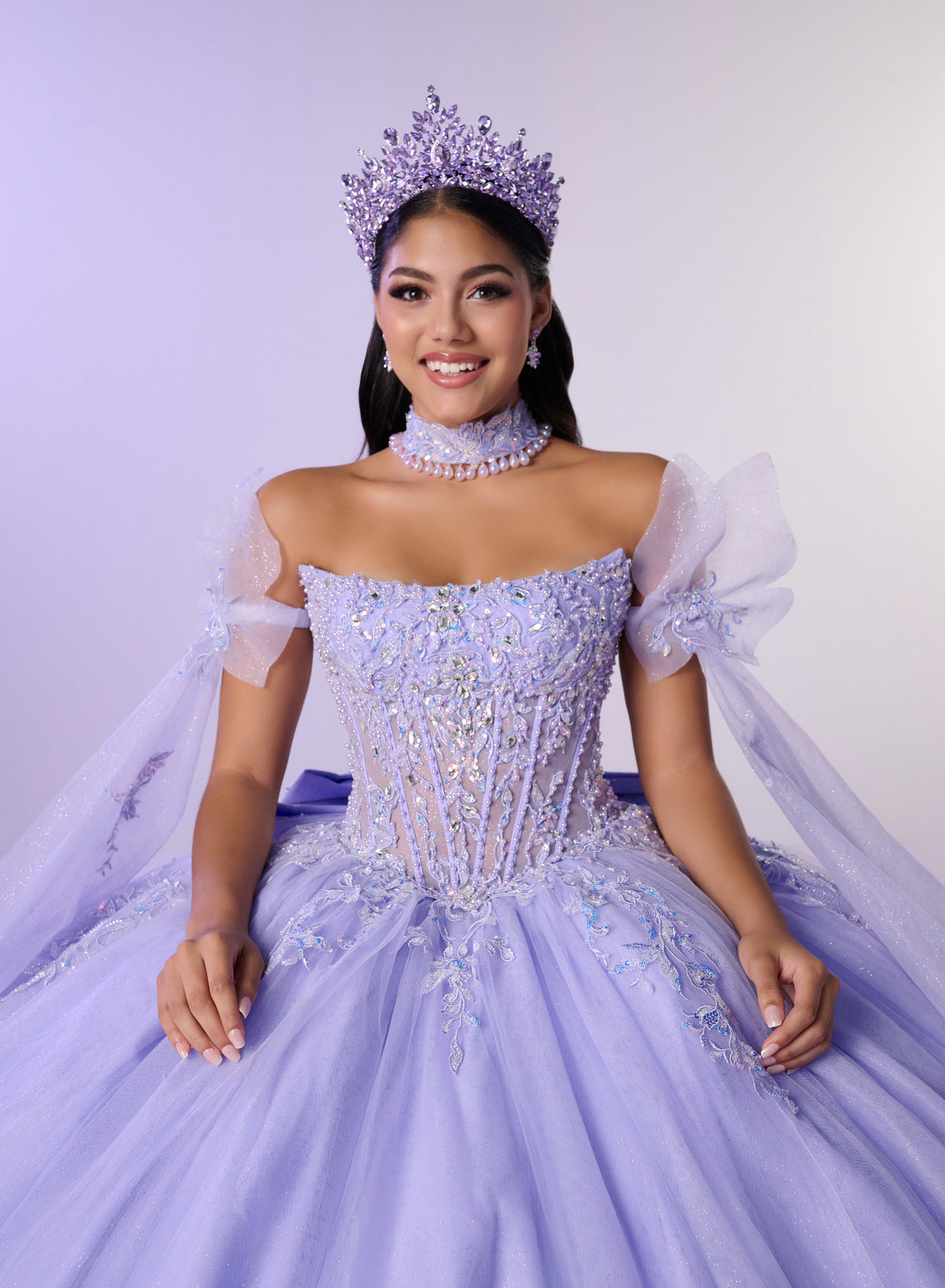 A woman wearing an elaborate lavender quinceañera-style ball gown with a jeweled corset bodice, sheer puff sleeves, a matching veil, tiara, and pearl choker, posed against a pale lavender studio background. / Dreamy off-shoulder ball gown featuring delicate sparkling appliqués, a structured corset bodice, a voluminous tulle skirt, and an oversized statement bow at the back.