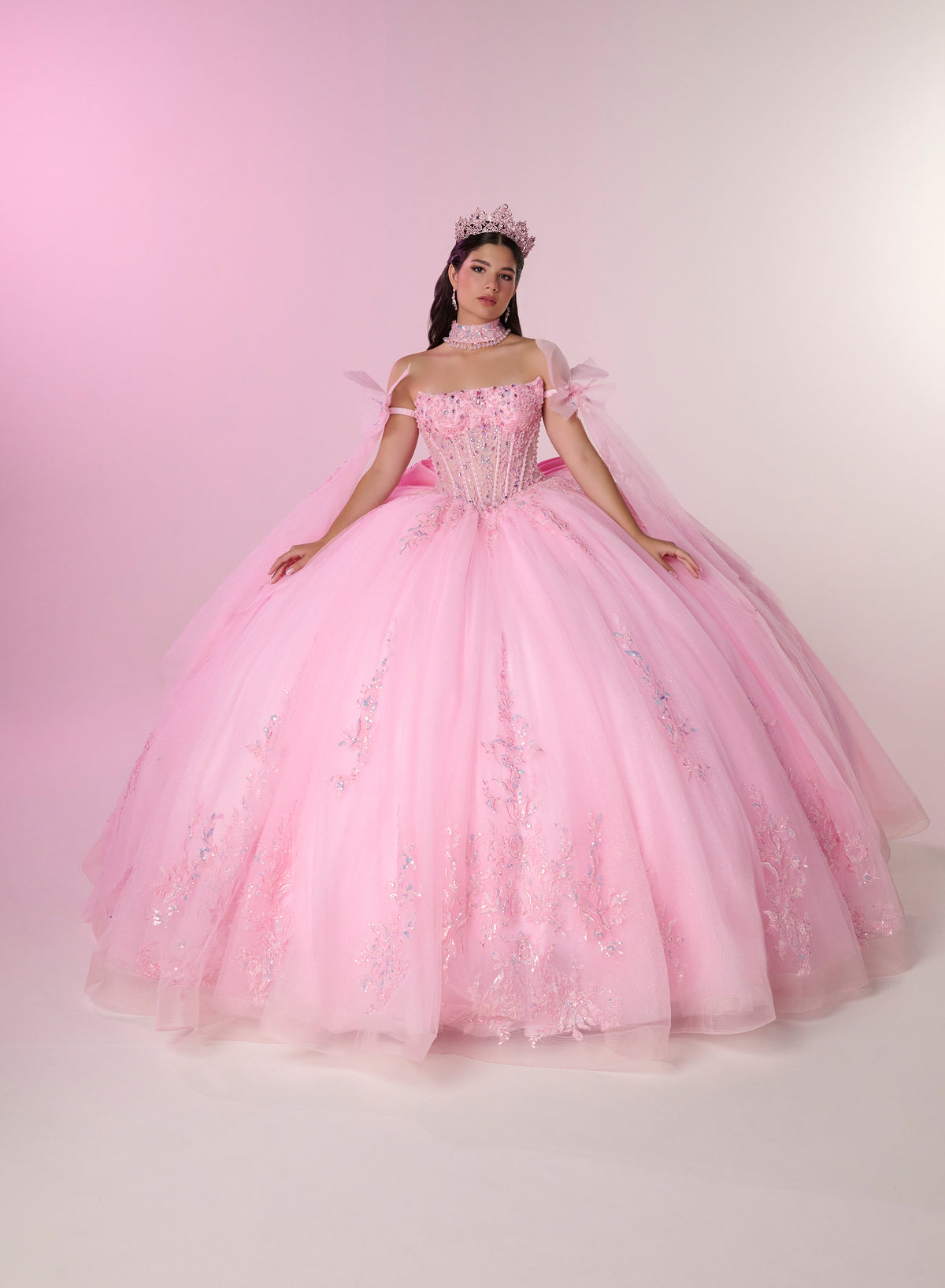 A young woman wearing a voluminous pink ball gown with floral embroidery and a matching pink tiara, posed against a soft pink studio backdrop. / Dreamy off-shoulder ball gown featuring delicate sparkling appliqués, a structured corset bodice, a voluminous tulle skirt, and an oversized statement bow at the back.