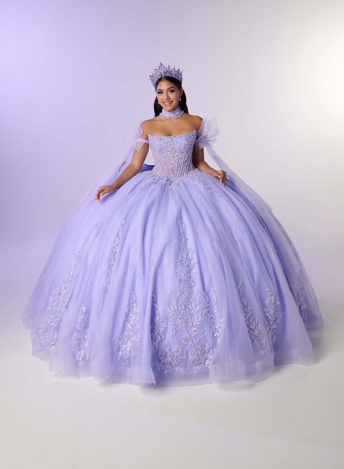 A woman modeling a voluminous lavender off-the-shoulder ball gown with lace embroidery, a matching headpiece, and a sheer cape, photographed against a light lavender studio backdrop. / Dreamy off-shoulder ball gown featuring delicate sparkling appliqués, a structured corset bodice, a voluminous tulle skirt, and an oversized statement bow at the back.