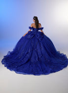 A woman wearing a voluminous royal blue ball gown, photographed from behind against a soft blue-to-white studio backdrop. The dress has off-the-shoulder ruffled sleeves, a fitted back with a large bow detail, and a full layered tulle skirt with floral embroidery. / Elegant off-shoulder ball gown adorned with intricate sparkling appliqués, a structured sweetheart bodice, a voluminous skirt, and a dramatic oversized bow with a sweeping train.
