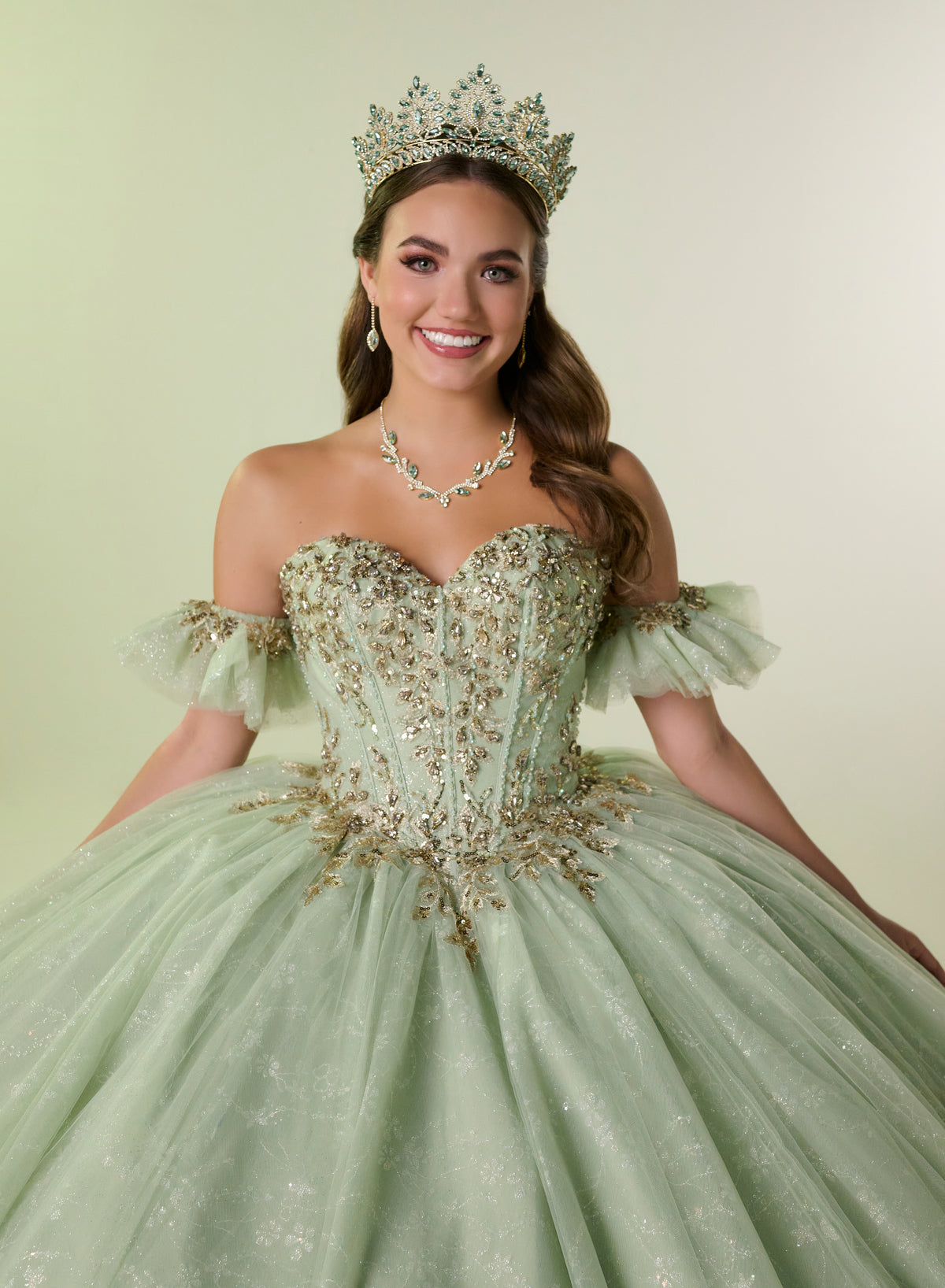 A smiling young woman wearing a mint green off-the-shoulder ball gown with gold beaded embroidery, a matching tiara, and a matching necklace, posed against a solid pale green studio background. / Elegant off-shoulder ball gown adorned with intricate sparkling appliqués, a structured sweetheart bodice, a voluminous skirt, and a dramatic oversized bow with a sweeping train.