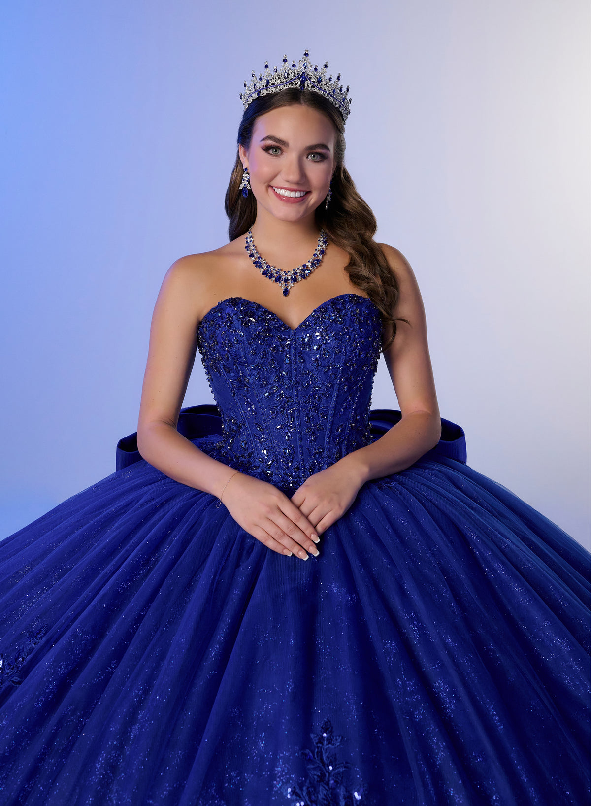 A young woman wearing a sparkling royal blue strapless ball gown with a sweetheart bodice, full tulle skirt, matching blue tiara, and blue beaded necklace, posed against a light blue studio background. / Elegant off-shoulder ball gown adorned with intricate sparkling appliqués, a structured sweetheart bodice, a voluminous skirt, and a dramatic oversized bow with a sweeping train.