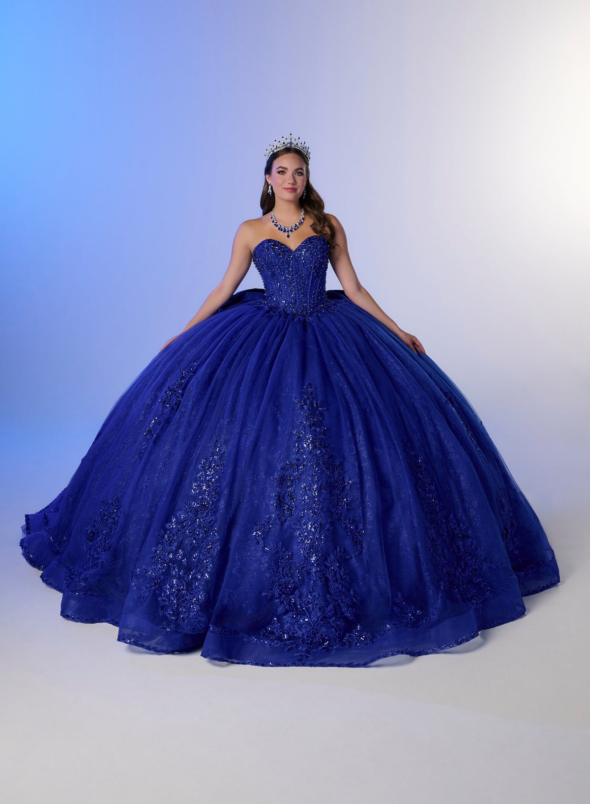 A woman wearing a voluminous royal blue ball gown with lace detailing and a tiara, posed against a blue-to-white studio backdrop. / Elegant off-shoulder ball gown adorned with intricate sparkling appliqués, a structured sweetheart bodice, a voluminous skirt, and a dramatic oversized bow with a sweeping train.