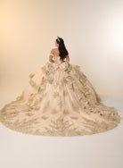 A woman viewed from behind wearing an ornate off-the-shoulder ball gown with a very full skirt, large bow detail at the back, and extensive floral embroidery and applique in a warm champagne-beige tone against a plain cream studio background. / Opulent off-shoulder ball gown featuring intricate embroidered appliqués, dramatic tiered draping, a structured corset bodice, and a sweeping cathedral-length train.