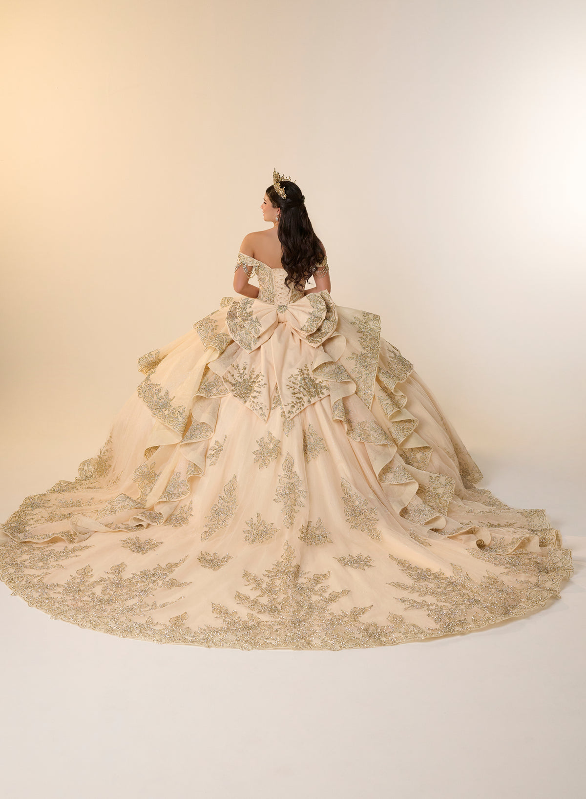 A woman viewed from behind wearing an ornate off-the-shoulder ball gown with a very full skirt, large bow detail at the back, and extensive floral embroidery and applique in a warm champagne-beige tone against a plain cream studio background. / Opulent off-shoulder ball gown featuring intricate embroidered appliqués, dramatic tiered draping, a structured corset bodice, and a sweeping cathedral-length train.