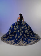 A woman seated on a voluminous royal blue ball gown with ornate gold floral embroidery, photographed from behind against a soft blue-to-gray studio backdrop. / Opulent off-shoulder ball gown featuring intricate embroidered appliqués, dramatic tiered draping, a structured corset bodice, and a sweeping cathedral-length train.