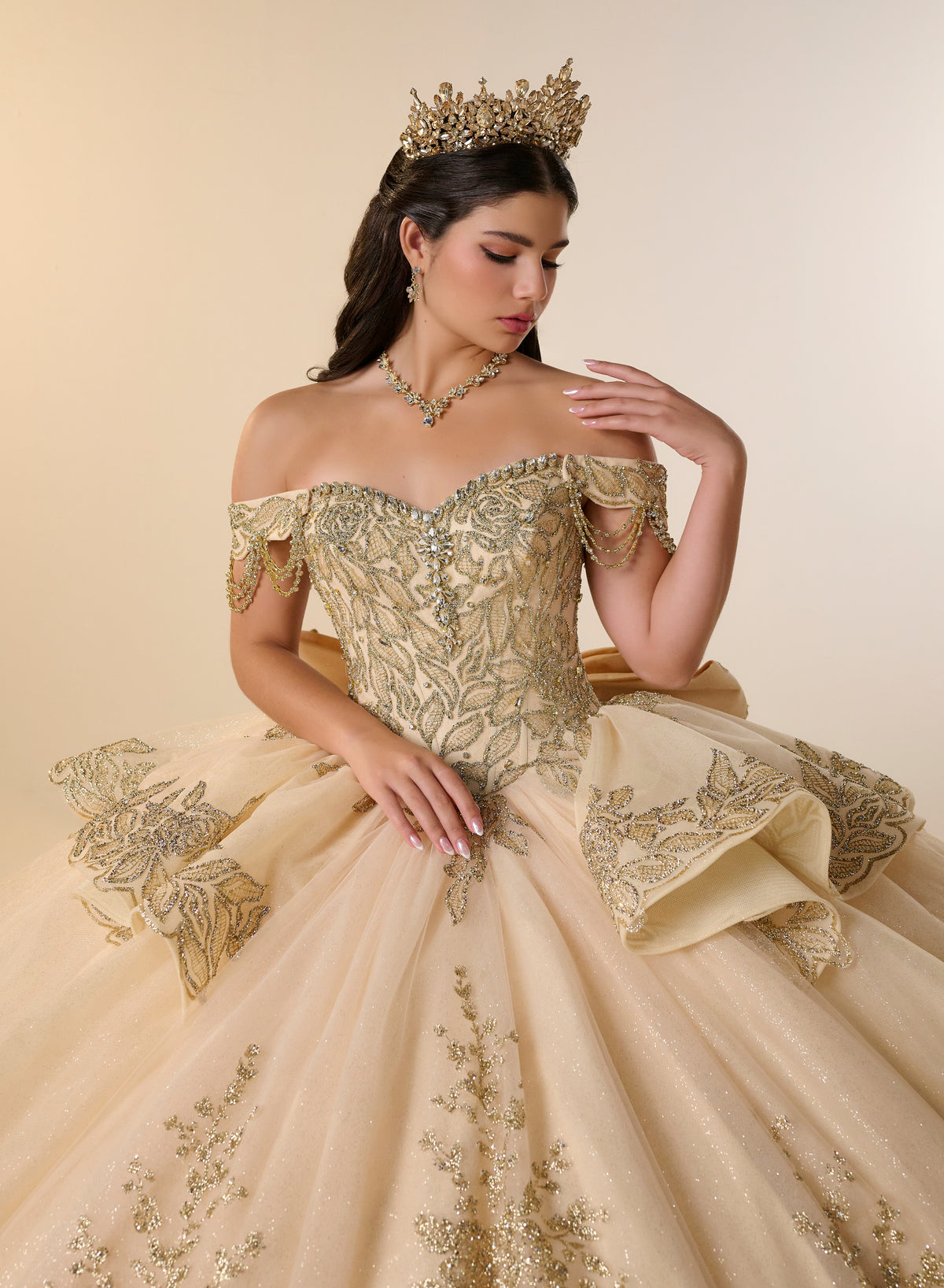 A woman wearing an ornate off-the-shoulder ball gown in champagne beige with intricate gold embroidery, a matching tiara, and a gold necklace, posed against a warm beige studio background. / Opulent off-shoulder ball gown featuring intricate embroidered appliqués, dramatic tiered draping, a structured corset bodice, and a sweeping cathedral-length train.