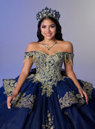 A woman wearing an ornate off-the-shoulder navy blue quinceañera gown with extensive gold embroidery, a matching crown, and a necklace, posed against a solid lavender-gray studio background. / Opulent off-shoulder ball gown featuring intricate embroidered appliqués, dramatic tiered draping, a structured corset bodice, and a sweeping cathedral-length train.