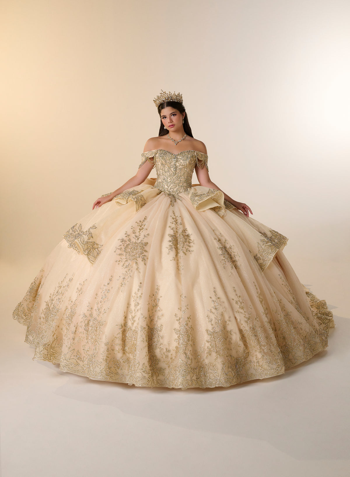 A woman modeling an elaborate off-the-shoulder ball gown in champagne beige with gold embroidery, a matching bow at the waist, and a gold crown, set against a plain beige studio background. / Opulent off-shoulder ball gown featuring intricate embroidered appliqués, dramatic tiered draping, a structured corset bodice, and a sweeping cathedral-length train.