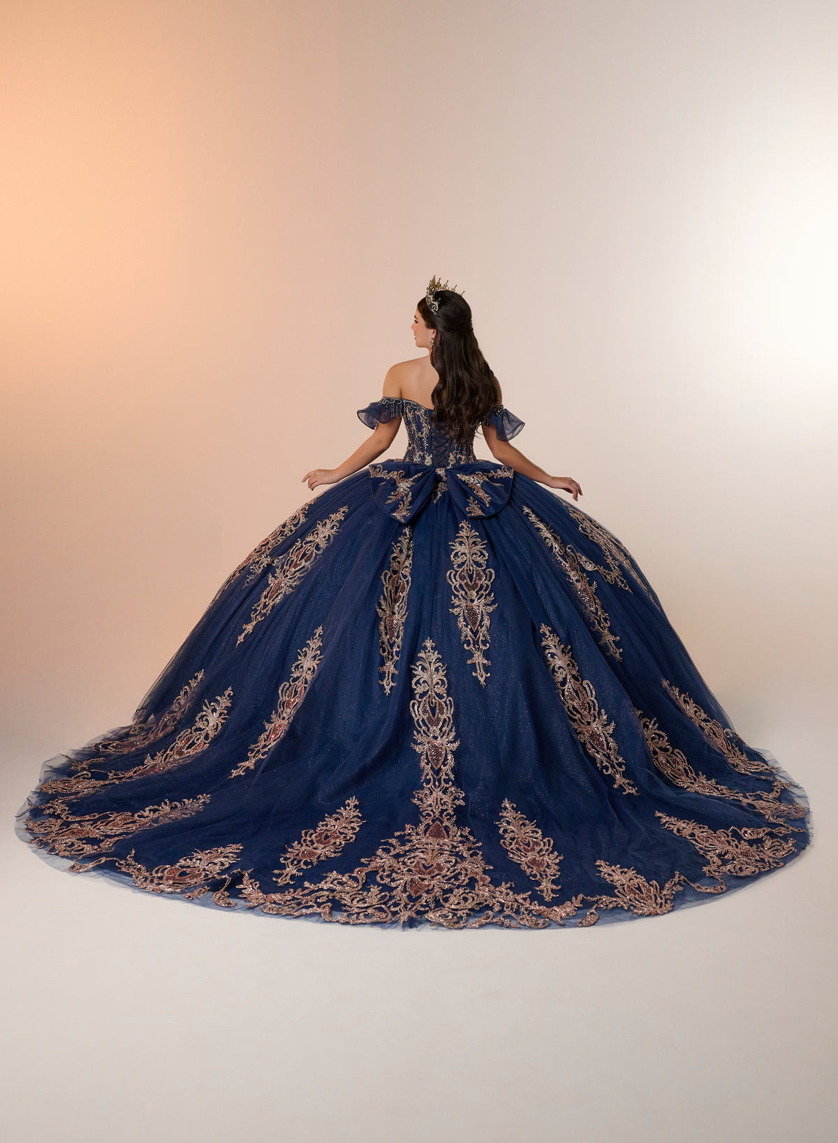 A woman viewed from behind wearing an elaborate off-the-shoulder ball gown in deep navy blue with ornate gold embroidery, set against a warm beige studio background. / Regal off-shoulder ball gown adorned with intricate embroidered appliqués, a structured corset bodice, and a voluminous skirt with an elegant sweeping train.