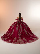 A woman viewed from behind wearing an elaborate off-the-shoulder ball gown in deep burgundy with ornate gold embroidery, set against a warm beige studio background. / Regal off-shoulder ball gown adorned with intricate embroidered appliqués, a structured corset bodice, and a voluminous skirt with an elegant sweeping train.