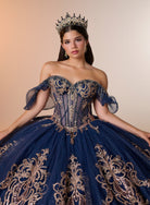 A woman wearing an elaborate off-the-shoulder ball gown in deep navy blue with ornate gold embroidery, a matching crown, and dangling earrings, posed against a warm beige studio background. / Regal off-shoulder ball gown adorned with intricate embroidered appliqués, a structured corset bodice, and a voluminous skirt with an elegant sweeping train.