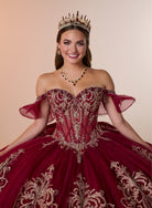 A smiling young woman wearing an ornate off-the-shoulder quinceañera dress in deep burgundy with intricate gold embroidery, a matching tiara, and a gold necklace, posed against a solid beige background. / Regal off-shoulder ball gown adorned with intricate embroidered appliqués, a structured corset bodice, and a voluminous skirt with an elegant sweeping train.