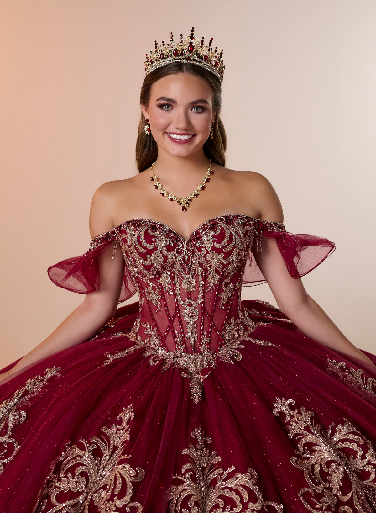 A smiling young woman wearing an ornate off-the-shoulder quinceañera dress in deep burgundy with intricate gold embroidery, a matching tiara, and a gold necklace, posed against a solid beige background. / Regal off-shoulder ball gown adorned with intricate embroidered appliqués, a structured corset bodice, and a voluminous skirt with an elegant sweeping train.