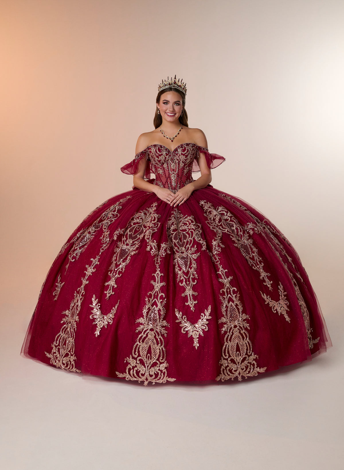 A woman wearing a voluminous off-the-shoulder quinceañera ball gown in deep burgundy with ornate gold embroidery, standing against a warm beige studio backdrop. She is also wearing a small tiara. / Regal off-shoulder ball gown adorned with intricate embroidered appliqués, a structured corset bodice, and a voluminous skirt with an elegant sweeping train.
