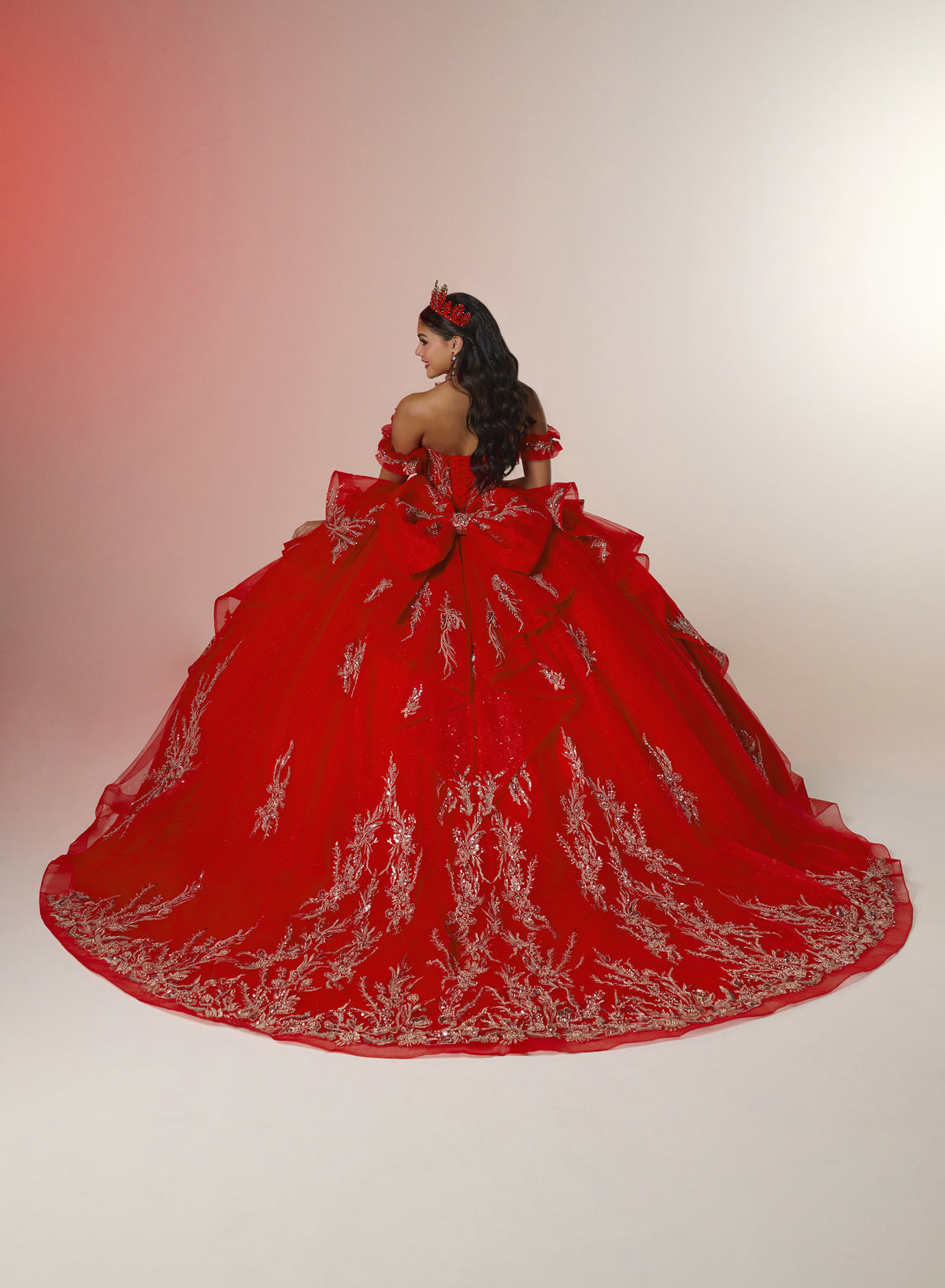 A woman wearing a voluminous red ball gown with silver floral embroidery, photographed from behind against a warm beige studio background. / Elegant off-shoulder ball gown featuring sparkling embellished appliqués, a structured corset bodice, dramatic layered skirt details, and a voluminous princess silhouette.