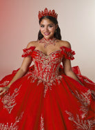 A woman wearing an ornate red quinceañera-style ball gown with silver embroidery, a matching red crown, and a red choker necklace, posed against a plain light beige studio background. / Elegant off-shoulder ball gown featuring sparkling embellished appliqués, a structured corset bodice, dramatic layered skirt details, and a voluminous princess silhouette.