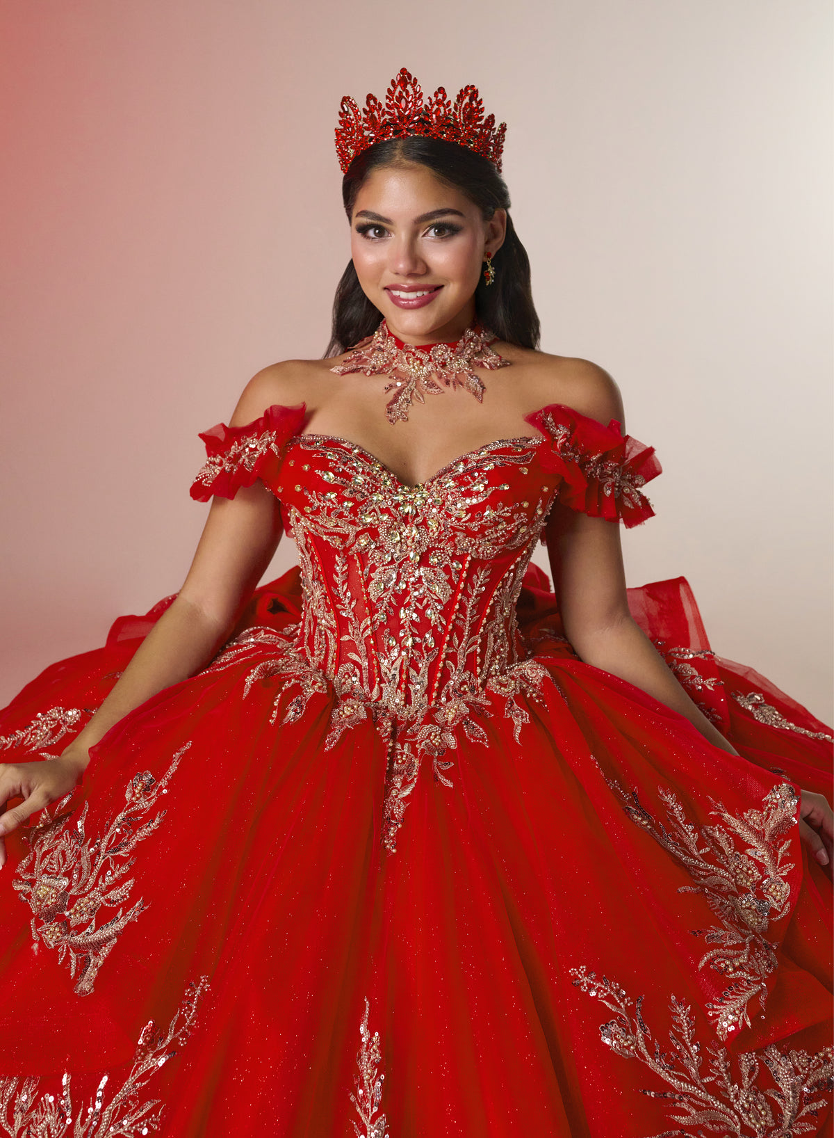 A woman wearing an ornate red quinceañera-style ball gown with silver embroidery, a matching red crown, and a red choker necklace, posed against a plain light beige studio background. / Elegant off-shoulder ball gown featuring sparkling embellished appliqués, a structured corset bodice, dramatic layered skirt details, and a voluminous princess silhouette.