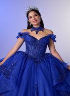 A woman wearing a royal blue off-the-shoulder ball gown with intricate beadwork, a matching blue choker, and a silver tiara, posing against a light blue studio background. / Elegant off-shoulder ball gown featuring sparkling embellished appliqués, a structured corset bodice, dramatic layered skirt details, and a voluminous princess silhouette.