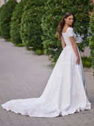 A woman wearing a white off-the-shoulder A-line wedding gown with a long train, standing on a paved walkway with green hedges in the background. / A-line style in jacquard with soft square neckline and short sleeves finished with scallop shape. Covered buttons at the back closure, buttons continuing all the way down to hemline along the train.
