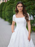A woman wearing a white A-line wedding dress with short puff sleeves, a square neckline, and a textured lace overlay, standing outdoors on a paved path with a green hedge in the background. / A-line style in jacquard with soft square neckline and short sleeves finished with scallop shape. Covered buttons at the back closure, buttons continuing all the way down to hemline along the train.