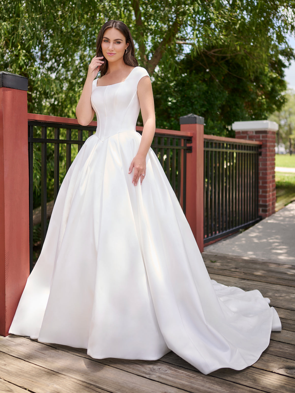 A woman wearing a white satin A-line wedding gown with a square neckline, cap sleeves, and a full skirt, standing on a wooden deck outdoors next to a red railing and green trees. / Clean mikado ball gown with structured bodice and princess skirt with pleats. The drop waist bodice with exposed boning has square neckline and cup sleeves. Covered buttons all the way to hemline on CB closure finishes the look.