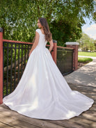 Woman in a white wedding dress standing on a wooden deck with greenery in the background / Clean mikado ball gown with structured bodice and princess skirt with pleats. The drop waist bodice with exposed boning has square neckline and cup sleeves. Covered buttons all the way to hemline on CB closure finishes the look.