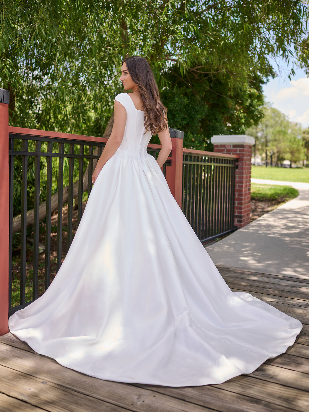 Woman in a white wedding dress standing on a wooden deck with greenery in the background / Clean mikado ball gown with structured bodice and princess skirt with pleats. The drop waist bodice with exposed boning has square neckline and cup sleeves. Covered buttons all the way to hemline on CB closure finishes the look.