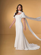 A woman modeling a floor-length ivory wedding gown with a high draped neckline, short flutter sleeves, and a long flowing train against a warm tan studio backdrop. / Slim crepe dress with square neckline and puffy short sleeves. Buttons at the back along zipper closure and all the way down on train.