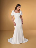 A woman modeling a floor-length ivory wedding gown with puff sleeves, a square neckline, and a slight train, posed against a warm tan studio backdrop. / Slim crepe dress with square neckline and puffy short sleeves. Buttons at the back along zipper closure and all the way down on train.
